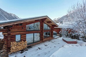 Une cabane en rondins dotée de grandes fenêtres, d’un toit recouvert de neige et d’une pile de bois de chauffage à l’extérieur.