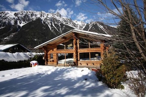 A wooden cabin with a snow-covered roof, surrounded by mountains.