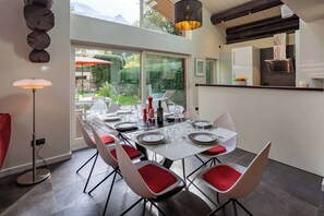 A modern dining area with a large table set for six, surrounded by white chairs with red cushions.
