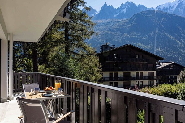 A balcony with a table set for breakfast, overlooking a mountain landscape and traditional chalet-style buildings.