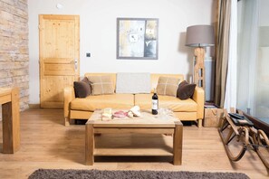 A cozy living room with a wooden floor, a yellow sofa with cushions, a wooden coffee table with food and wine, a lamp, and a stone wall.