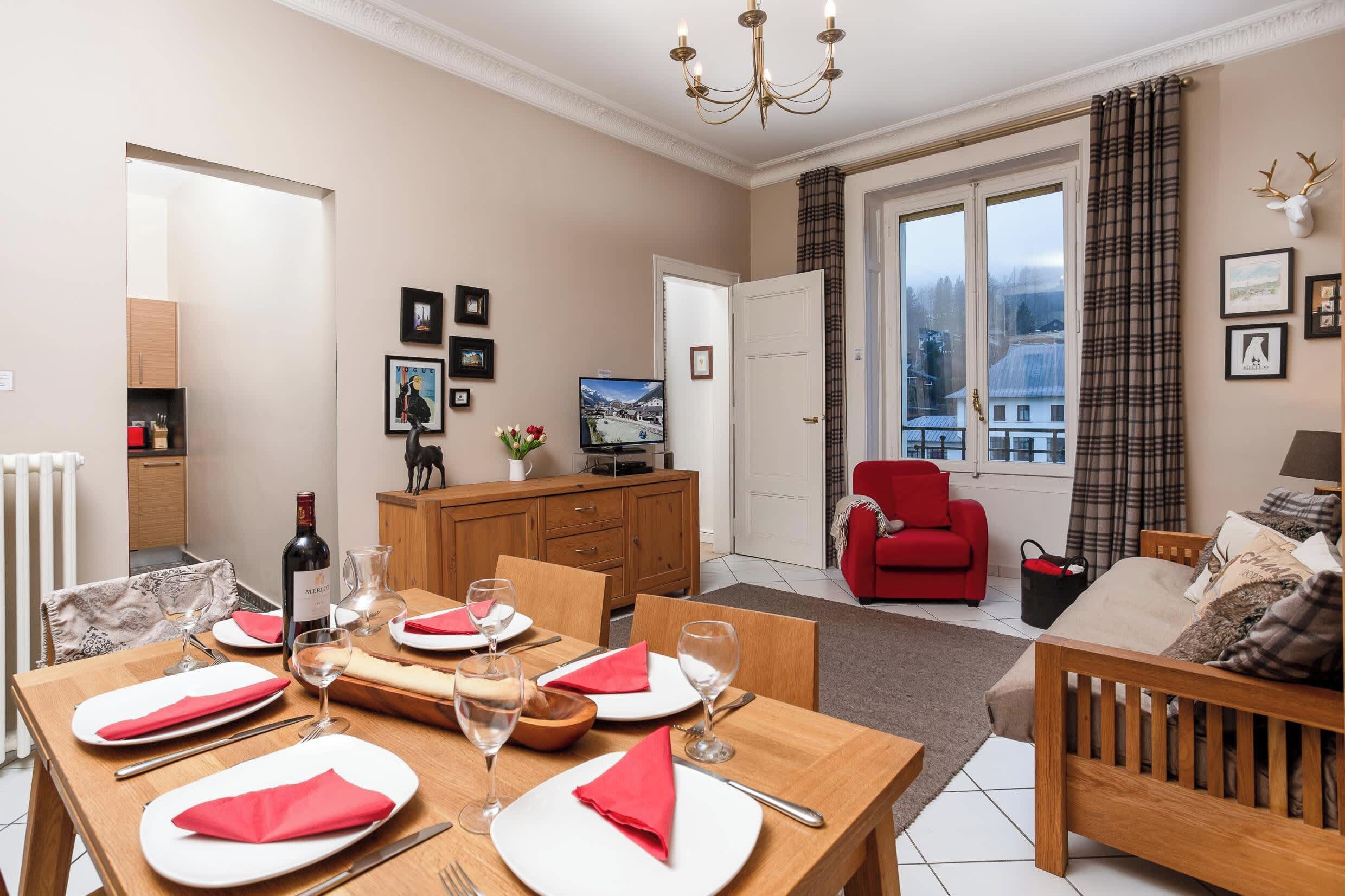 A dining area with a wooden table set for four, a television in the background, and a red armchair.