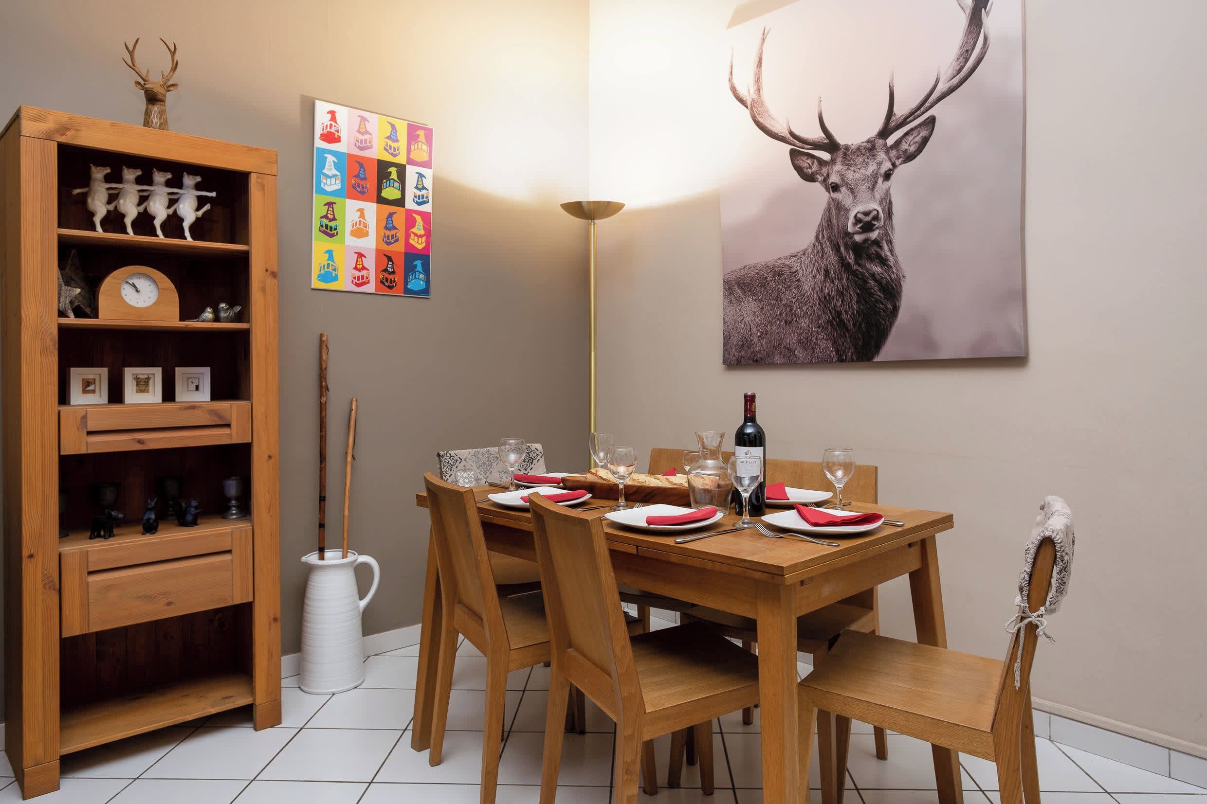 A dining area with a wooden table set for four, surrounded by chairs. A wooden cabinet with decorative items is to the left, and a colorful board game is on the wall.
