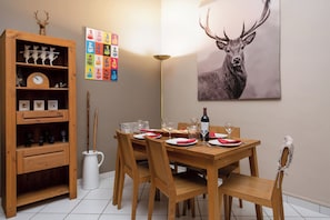 A dining area with a wooden table set for four, surrounded by chairs. A wooden cabinet with decorative items is to the left, and a colorful board game is on the wall.