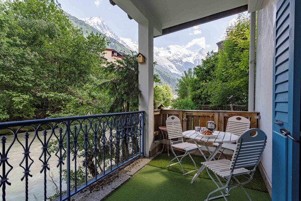 A balcony with a view of a river and mountains, a table with chairs, and a wooden deck.