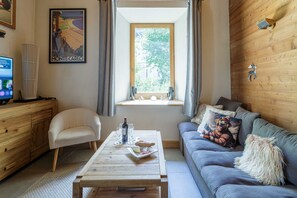 A cozy living room with a wooden floor, a grey sofa, a wooden coffee table with a bottle and glasses, a white armchair, and a window with curtains.