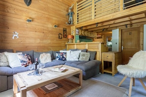 A cozy cabin interior with a wooden ceiling, a sofa, a coffee table, a fluffy chair, and a kitchen area.