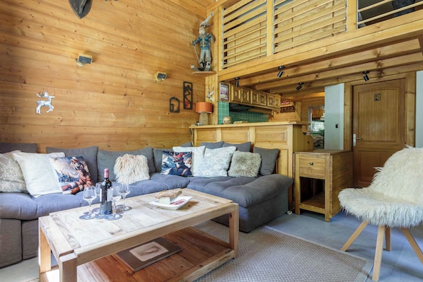 A cozy cabin interior with a wooden ceiling, a grey sofa, a wooden coffee table, and a white furry chair.