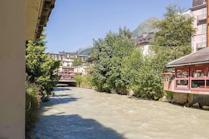 Une rivière étroite, bordée de bâtiments de chaque côté, un pont visible au loin et des montagnes en arrière-plan.