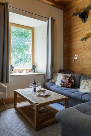 A cozy room with a wooden ceiling, a sofa with cushions, a window with curtains, and a table with glasses and a bottle. - Lou Lou Apartment -  an apartment that sleeps 4 guests  in 1 bedroom (Chamonix)