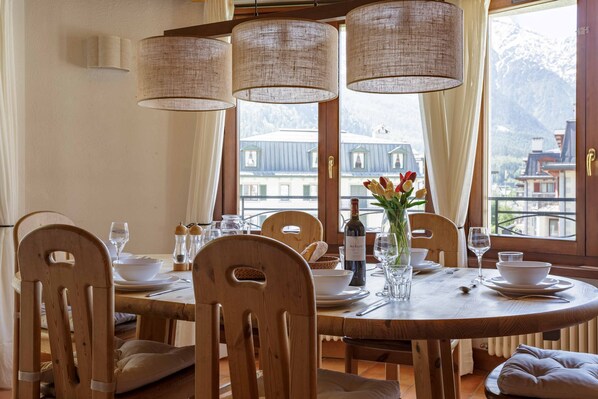 A dining area with a round wooden table, white chairs, and a view of mountains through large windows.