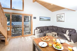A modern living room with a wooden staircase, a gray sectional sofa, a dining table with breakfast, and a large window with a mountain view. - Sauberands 18 apartment, Skiing, Chamonix All Year (Chamonix)