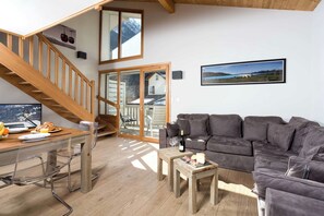 A spacious living area with a wooden staircase, a grey sofa, a dining table with breakfast items, and a large window with a mountain view.