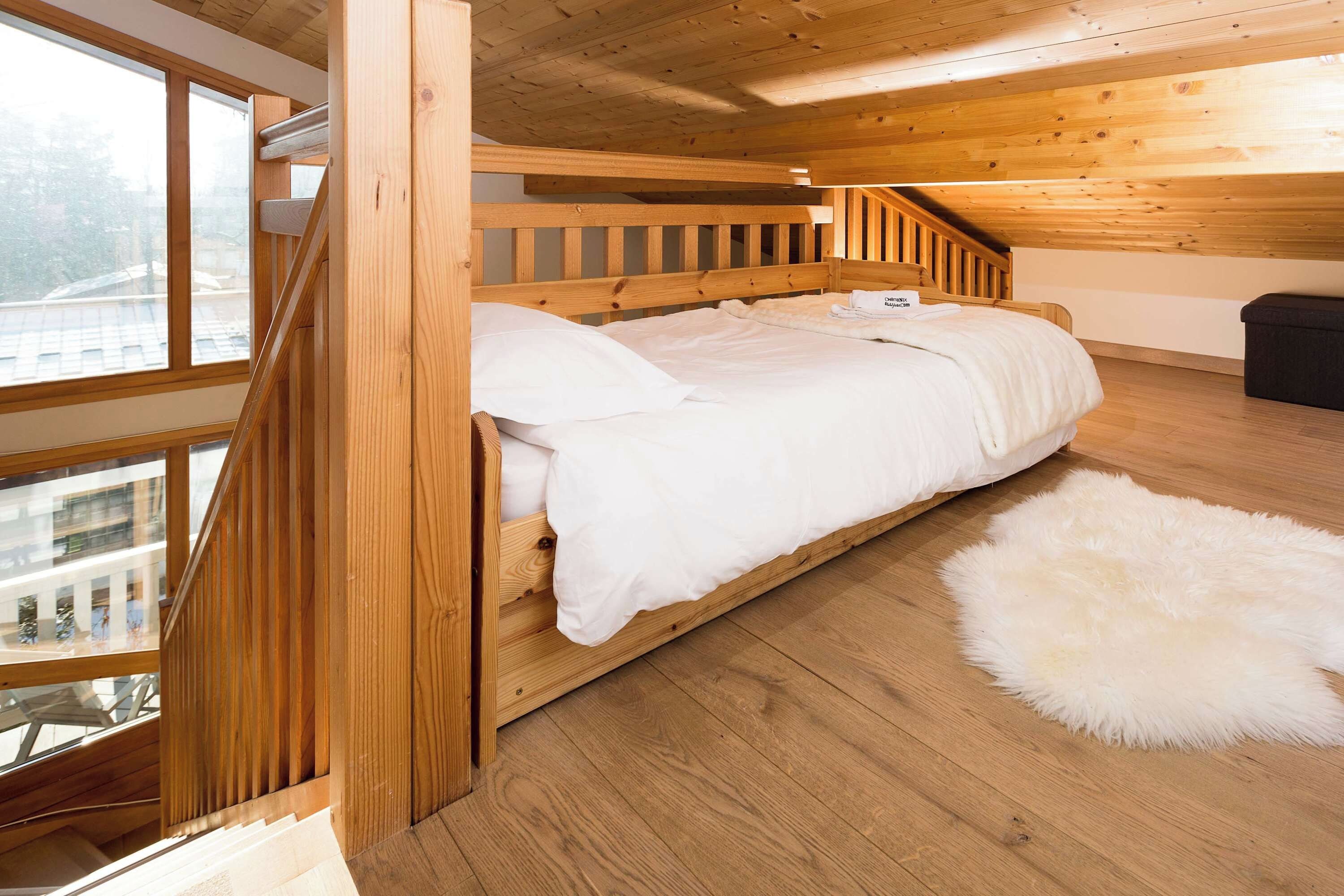 A wooden bunk bed with a white mattress and pillows, a fluffy white rug, and a window with a view of buildings.