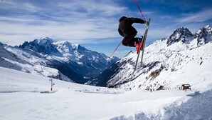 A skier mid-air performing a jump on a snowy mountain slope with a clear sky and snow-covered peaks in the background.