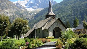 A stone church with a steeply pitched roof and a cross on top, surrounded by lush greenery and a mountain backdrop.