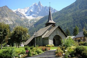 A stone church with a steeply pitched roof and a cross on top, surrounded by lush greenery and a mountain backdrop. - La Ferme des Praz appt, Golfing, Chamonix All Year (Chamonix)