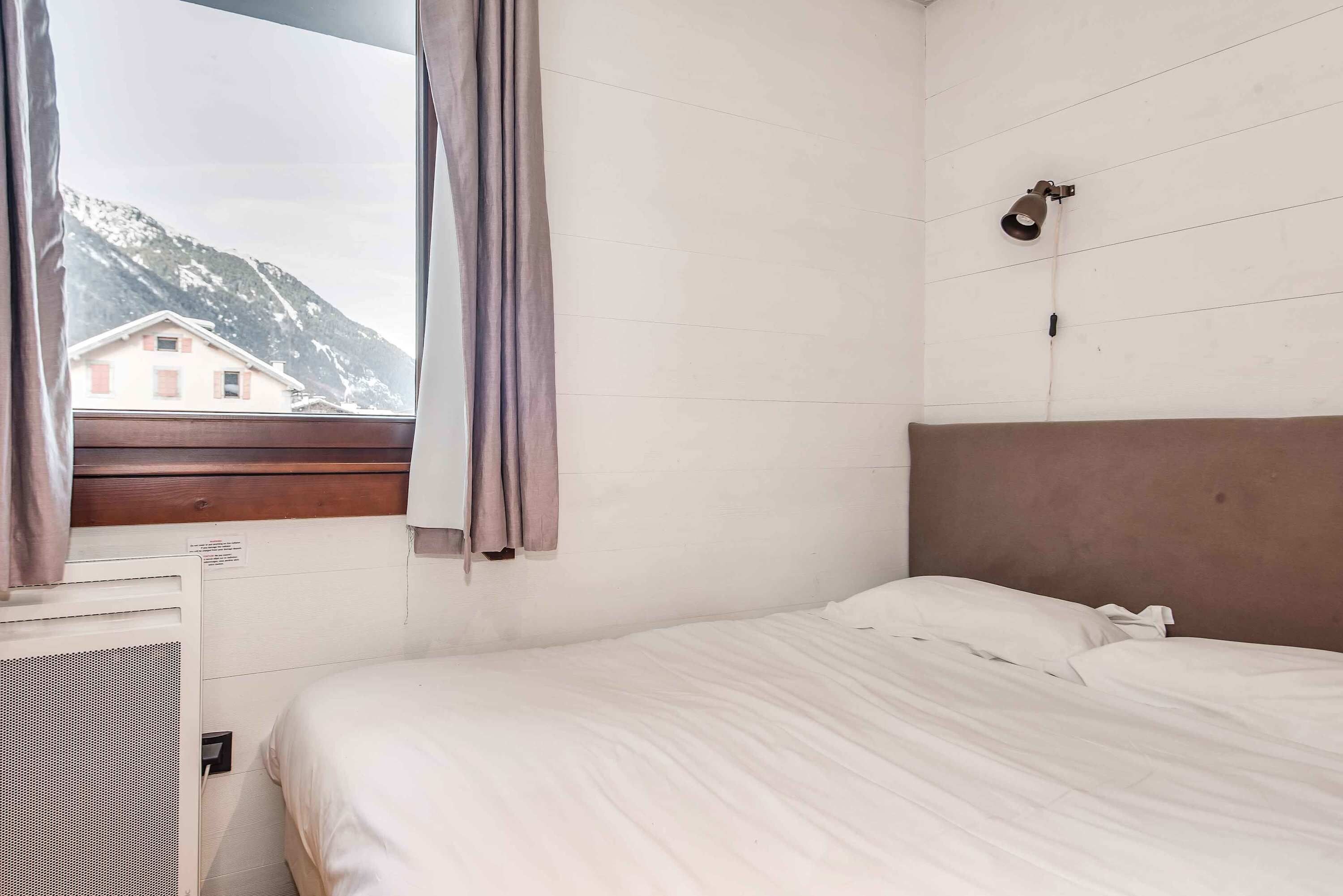 A hotel room with a bed, a window with curtains, a wall-mounted light, and a view of a mountain.