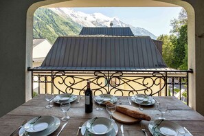 A dining table set for four with a bottle of wine, glasses, plates, and cutlery. The table is outdoors with a view of a mountain and a building with a metal roof.
