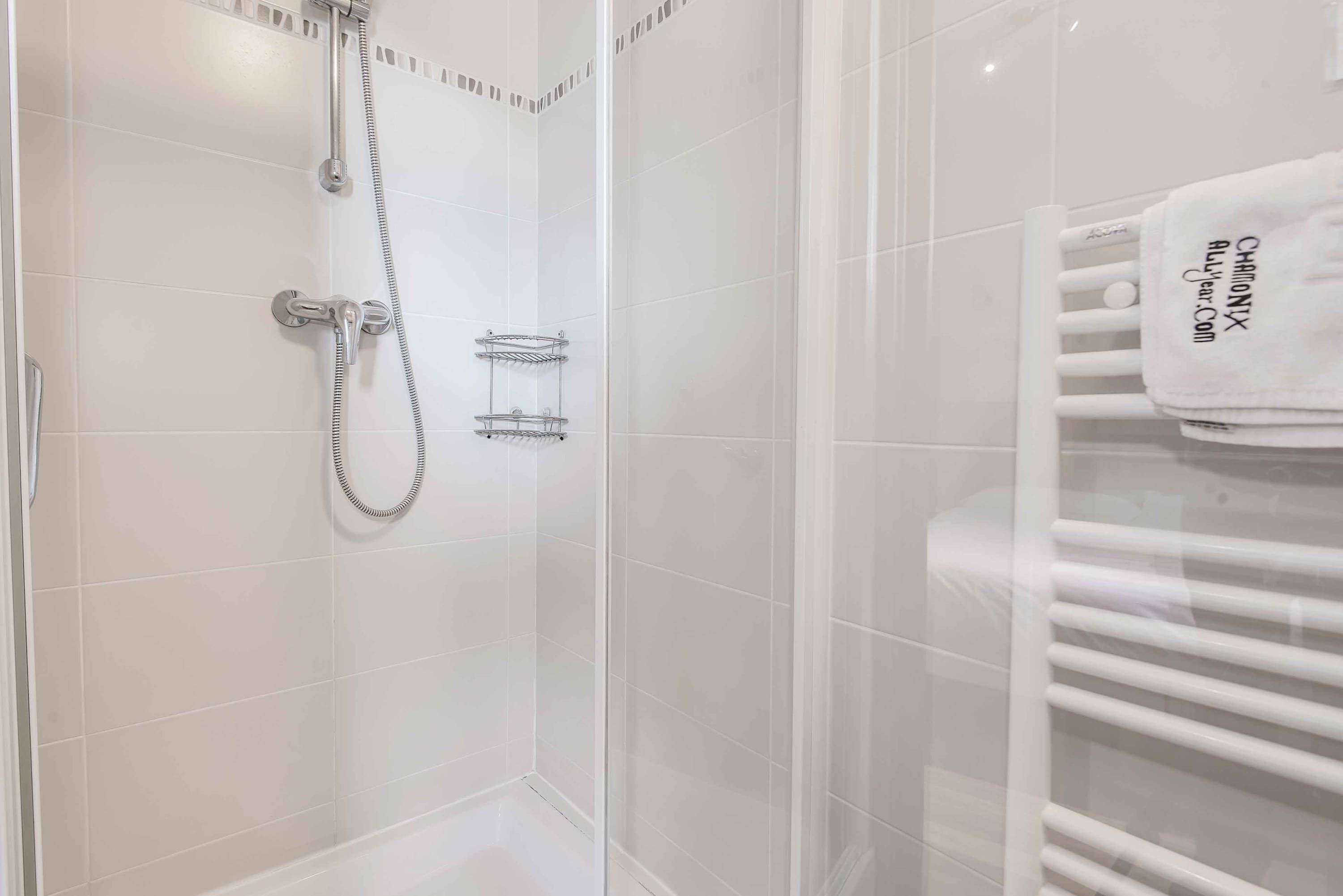 Un baño con azulejos blancos, una ducha con cabezal de ducha de mano y un perchero con una toalla blanca.