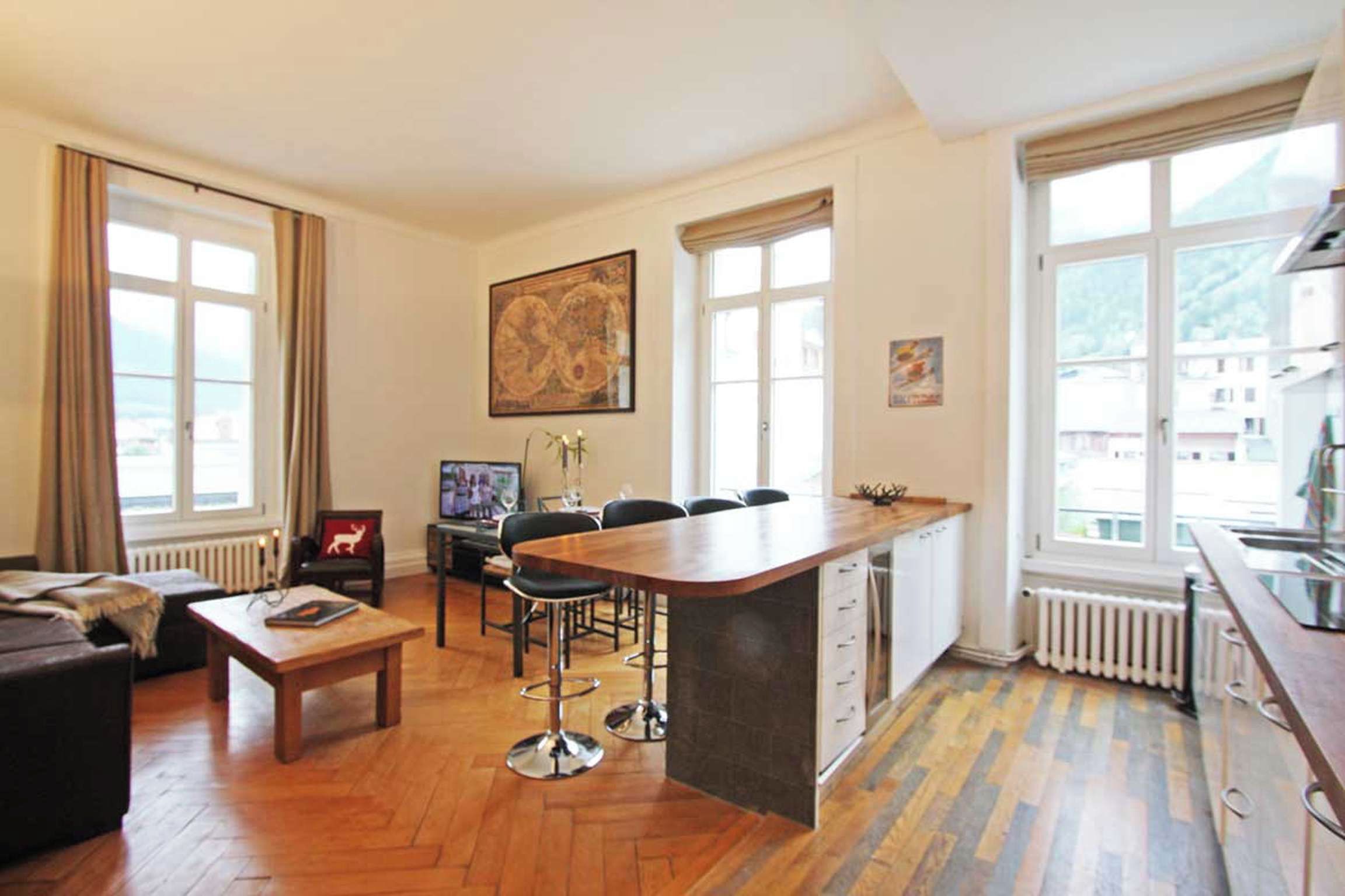 Een moderne woonkamer met een houten vloer, een bank, een salontafel, en een eethoek met stoelen. Er is een keuken met een bar en een groot raam.