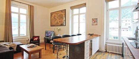 Een moderne woonkamer met een houten vloer, een bank, een salontafel, en een eethoek met stoelen. Er is een keuken met een bar en een groot raam.