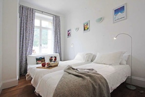 Une chambre à coucher avec un lit, une table de chevet avec une lampe, une fenêtre avec des rideaux et des œuvres d’art murales.