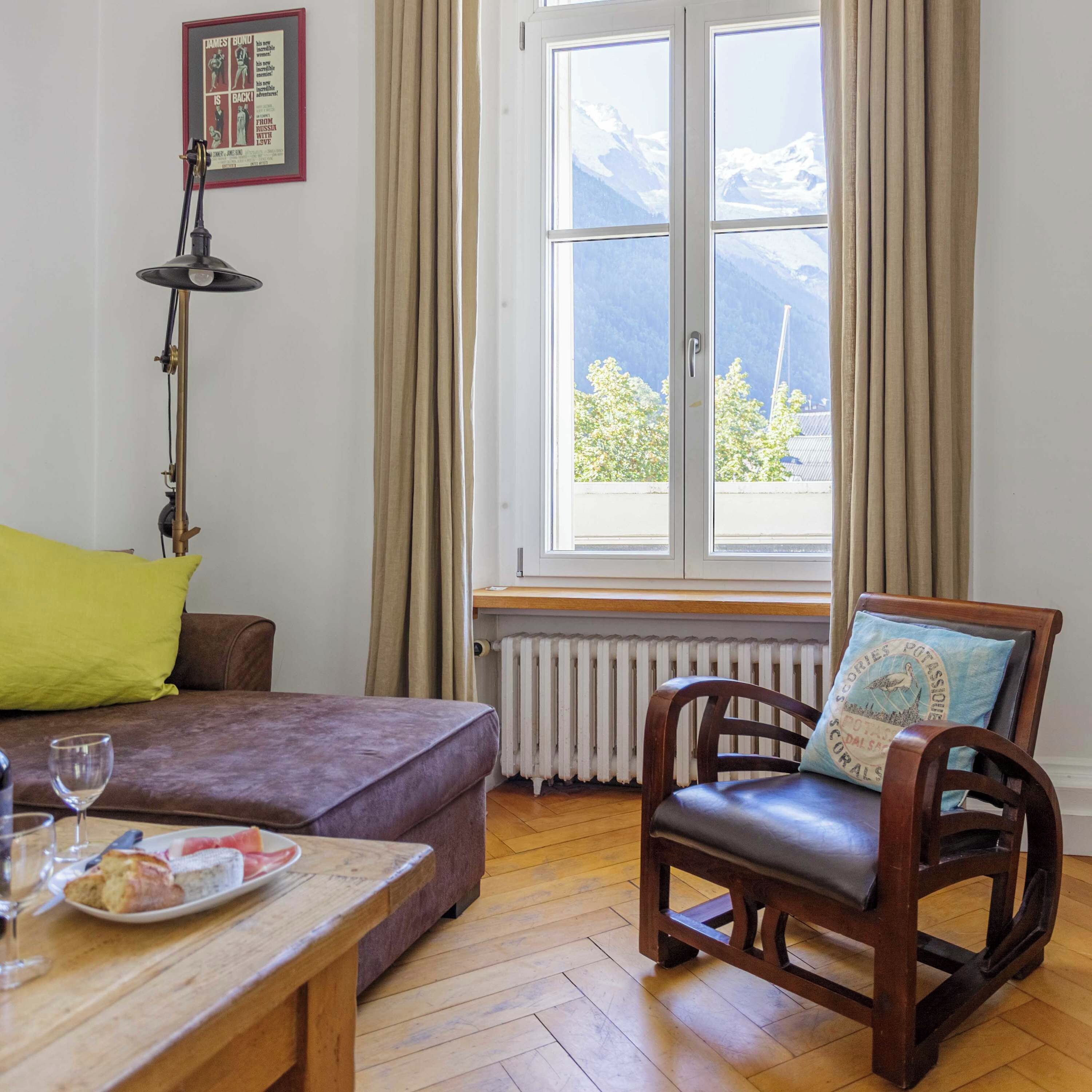 Une pièce comprenant un canapé, une chaise en bois, une table avec une assiette de nourriture, un verre à vin, une lampe, une fenêtre avec des rideaux et un radiateur.
