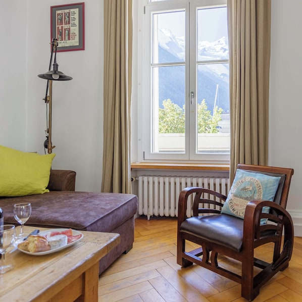 Une pièce comprenant un canapé, une chaise en bois, une table avec une assiette de nourriture, un verre à vin, une lampe, une fenêtre avec des rideaux et un radiateur.