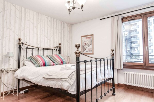 A bedroom with a metal bed frame, a white bedspread, and striped pillows. There is a window with a view of buildings, a radiator, and a poster on the wall.