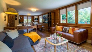 Un salon chaleureux avec un canapé, une table basse sur laquelle sont posées des boissons, et une vue sur la cuisine et le jardin grâce à de grandes fenêtres.