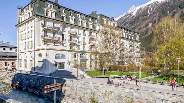A historic hotel with multiple balconies, a stone facade, and a view of a mountain range.
