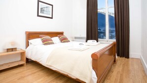 A neatly made bed with a wooden headboard, a bedside table with a lamp, and a window with curtains.