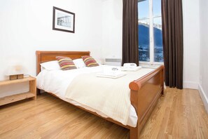 A neatly made bed with a wooden headboard, a bedside table with a lamp, and a window with curtains. - Historical Mont Blanc 98 Apt - Chamonix All Year (Chamonix)