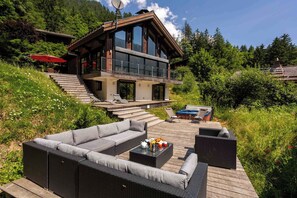 Une maison moderne à deux étages, dotée d’une terrasse, de mobilier d’extérieur et offrant une vue sur la forêt environnante.