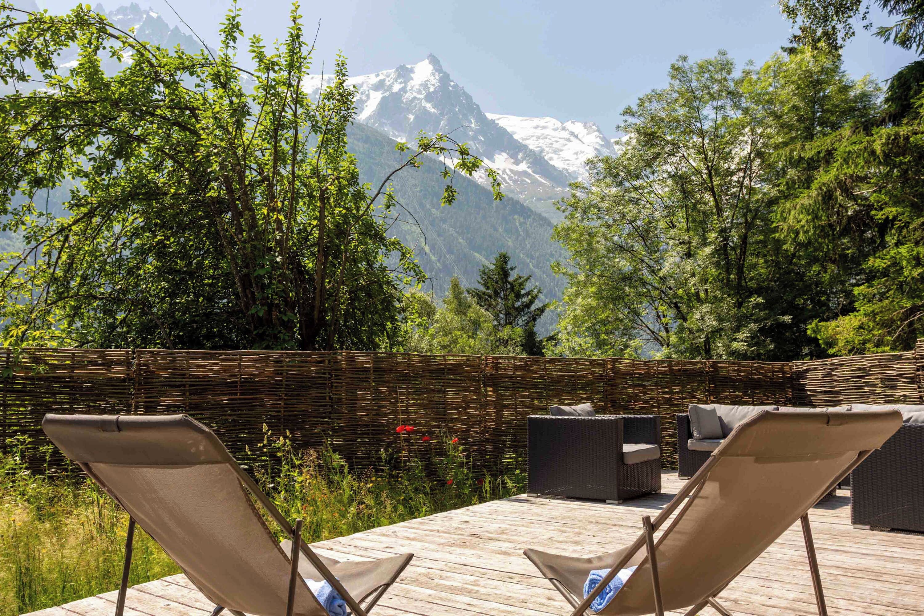 Une terrasse en bois, dotée de chaises longues et d’une clôture en osier, offre une vue sur un paysage montagneux.