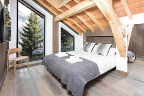 A modern bedroom with a large bed, a wooden chair, and a view of trees through the windows.