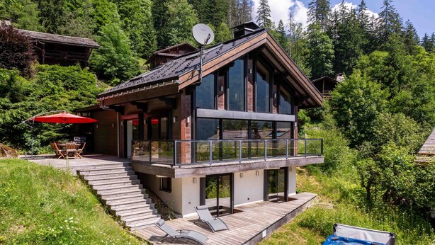Una moderna cabaña con terraza de madera, jacuzzi y zona de comedor con una sombrilla roja.