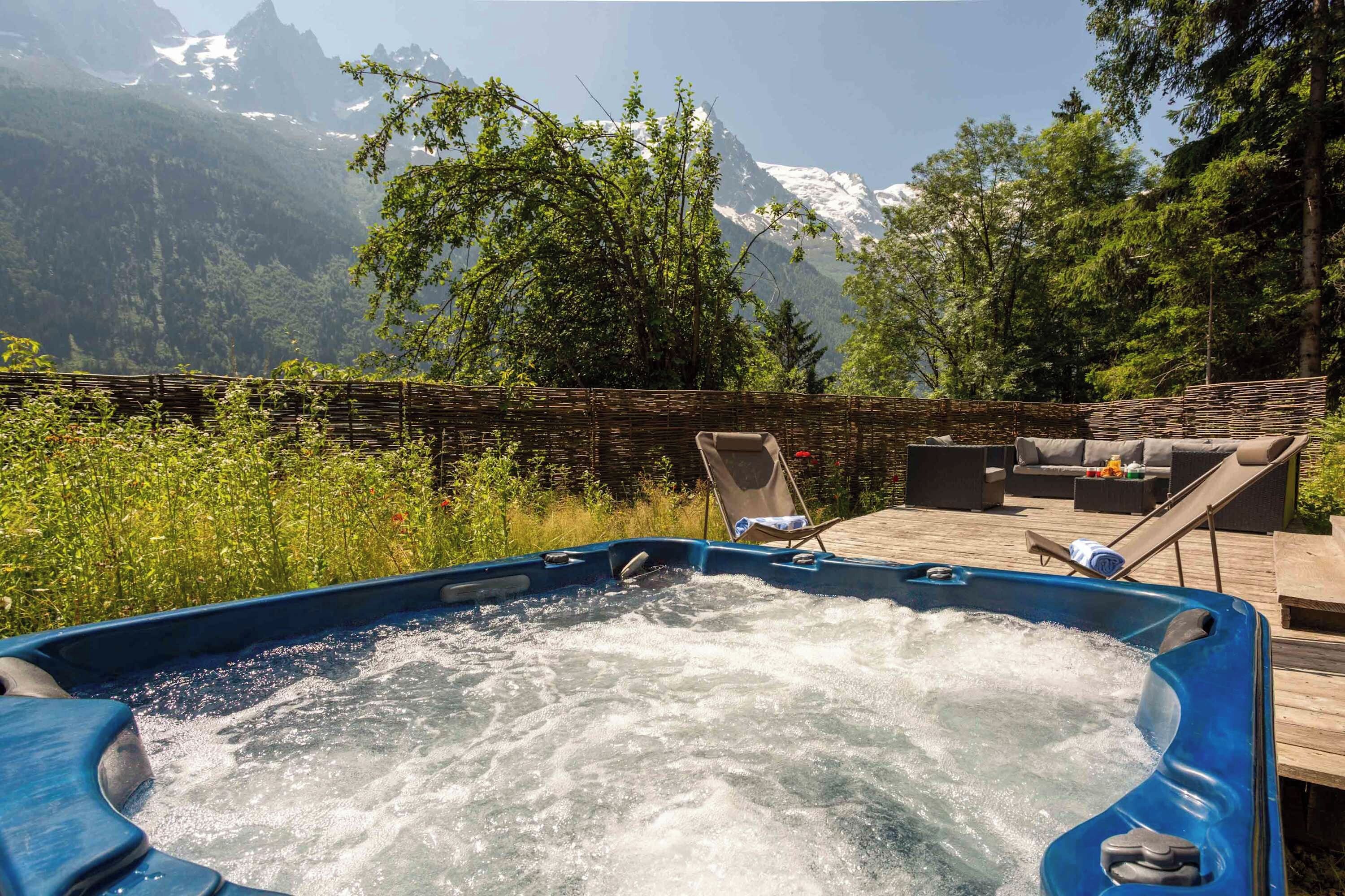 A hot tub with a wooden deck and outdoor furniture, surrounded by lush greenery and mountains in the background.