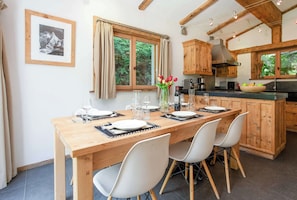 Un coin repas avec une table en bois dressée pour deux personnes, des chaises blanches, un vase de fleurs et, au fond, une cuisine.