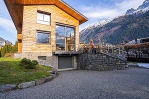 A modern log cabin with a stone foundation, large windows, and a stone staircase leading to a balcony.