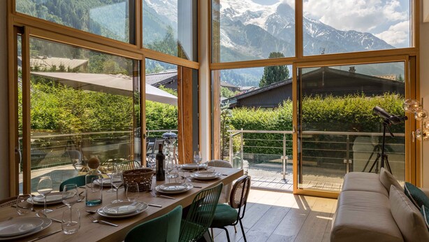 A dining area with a table set for four, large windows offering a mountain view, and a sofa in the foreground. - Chalet Couttet -  a self catered chalet that sleeps 8 guests  in 4 bedrooms (Chamonix)