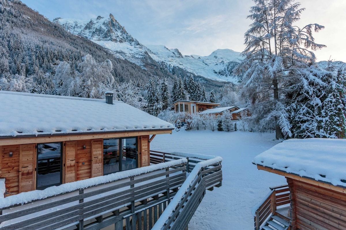 A wooden cabin with a balcony, surrounded by snow-covered trees and mountains.