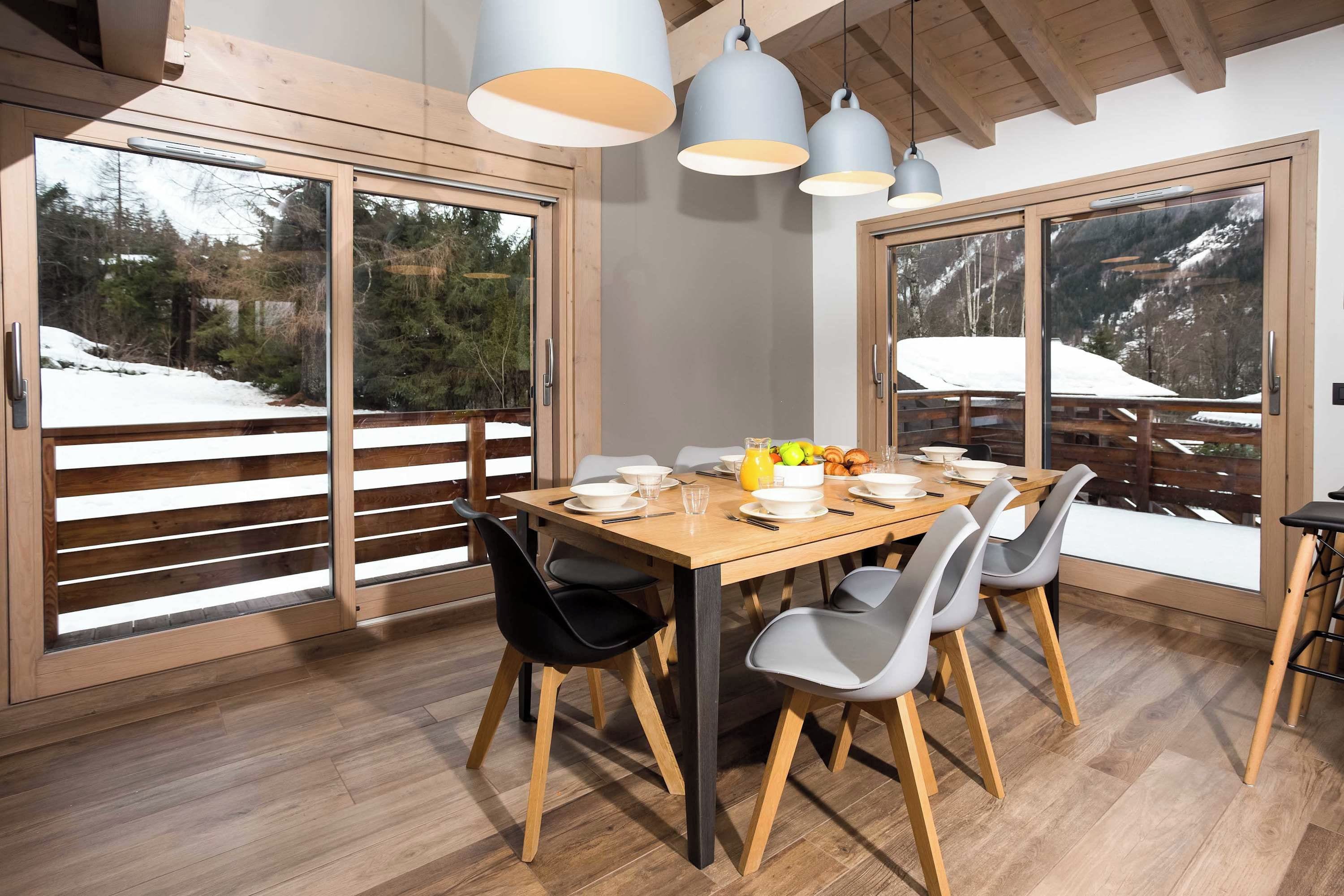 Un comedor con mesa de madera, sillas y vista a un paisaje nevado a través de amplios ventanales correderas.