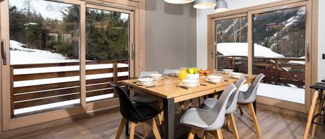 Un comedor con mesa de madera, sillas y vista a un paisaje nevado a través de amplios ventanales correderas.