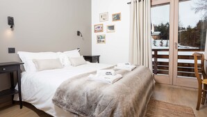 Een net opgemaakt bed met witte lakens, een zachte grijze plaid en keurig gevouwen handdoeken op een houten vloer.