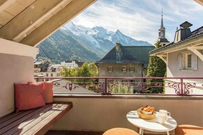 A balcony with a view of snow-capped mountains, a bench, and a table with a basket of bread and a cup.