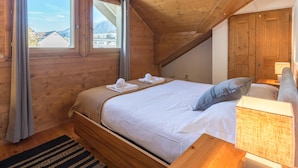 A bedroom with a wooden ceiling, a bed with white linens, a window with curtains, and a nightstand with a lamp.