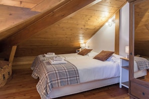 A cozy attic bedroom with a sloped ceiling, wooden beams, a bed with white linens, two pillows, a bedside table with a lamp, and a wicker basket. - Villa Vallet Apartment, Skiing, Chamonix All Year (Mont Blanc)