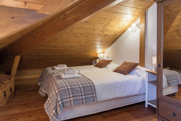 A cozy attic bedroom with a sloped ceiling, wooden beams, a bed with white linens, two pillows, a bedside table with a lamp, and a wicker basket.